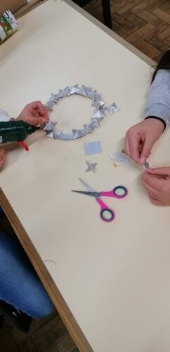 Uma aluna a construir uma coroa colando com cola quente, várias mini estrelas que outra aluna está a fazer.